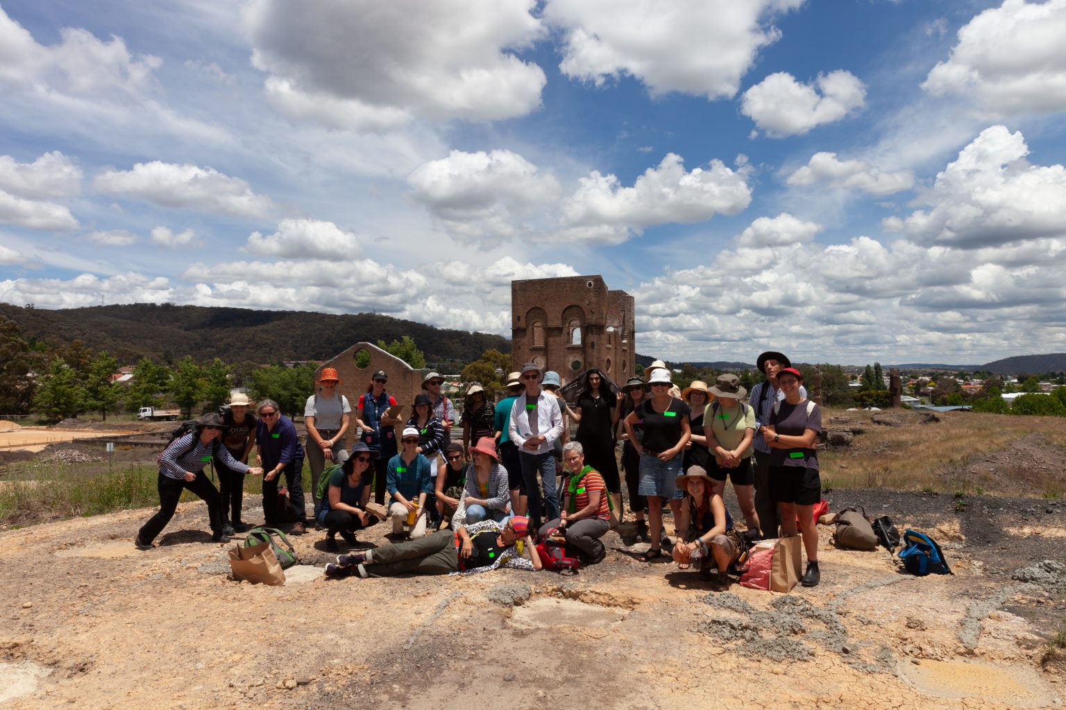 a group of about 20 people posing as a group in a rural town with a tall ruined industrial building in the background. There are white puffy clouds in the sky and it looks like a hot day