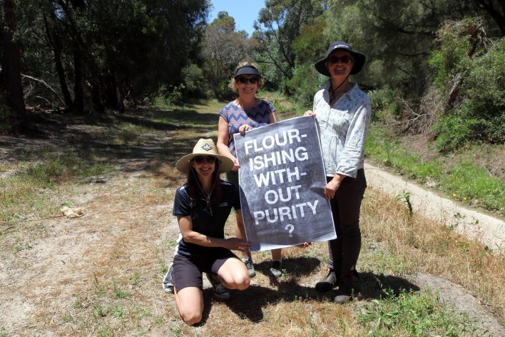 three women are gathered in the middle of an open trail with a dry drainage channel on the right hand side background. The three women are smiling in the sun and hold a black and white poster that says "flourishing without purity"