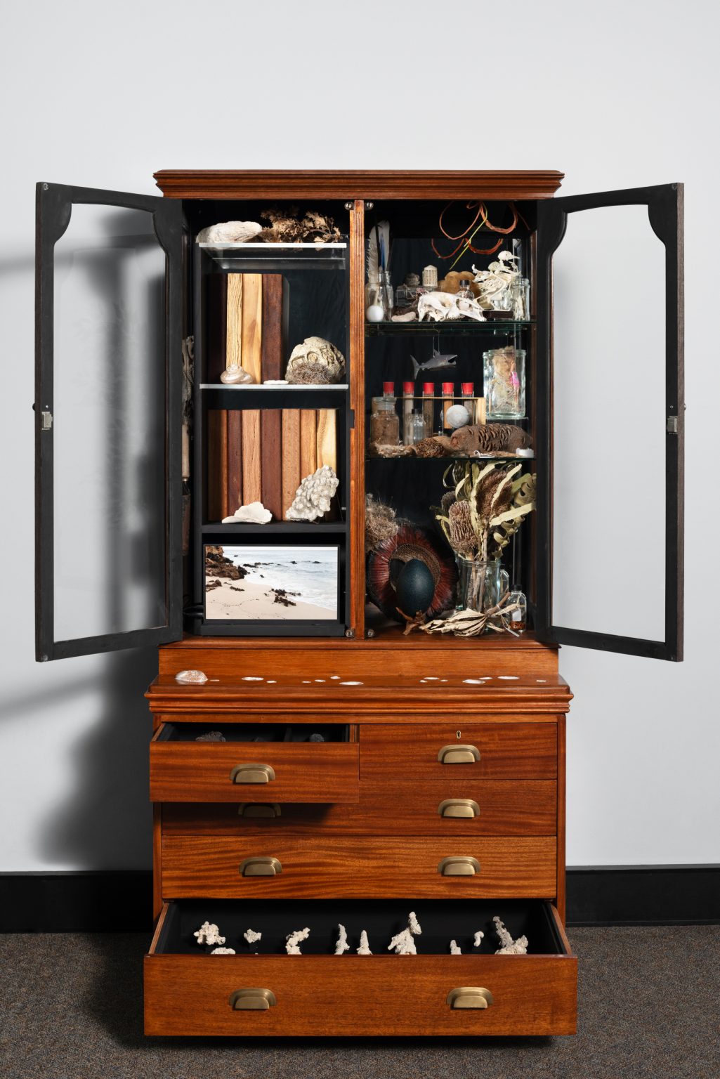 a tall wooden cupboard combining a glass cabinet and 5 drawers filled with many objects