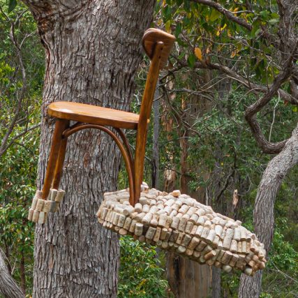 a wooden bentwood chair is suspended high in the forest canopy. It has a cloud of corks attached to the back legs. The image is tightly cropped