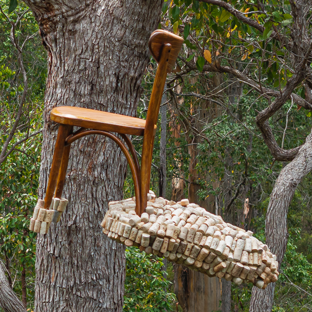a wooden bentwood chair is suspended high in the forest canopy. It has a cloud of corks attached to the back legs. The image is tightly cropped